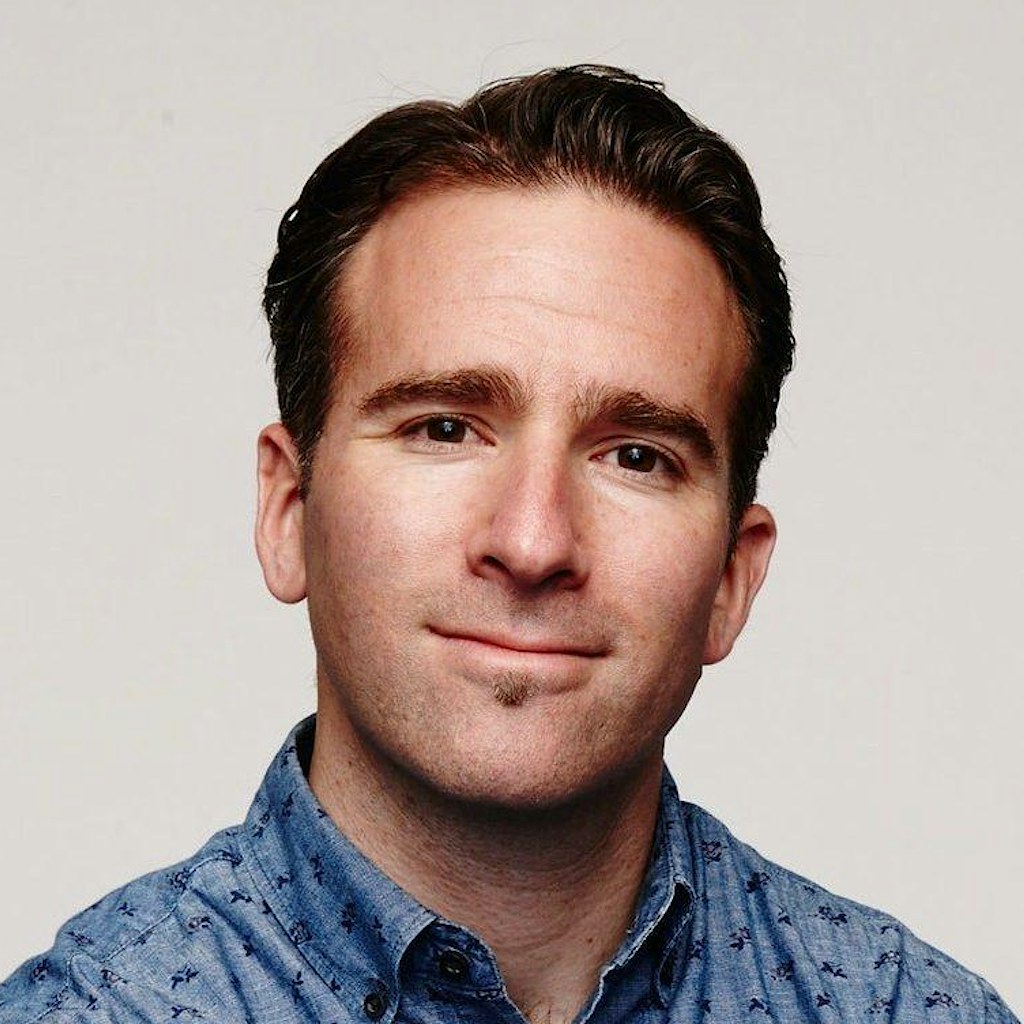 White man with blue button-down shirt looking at the camera. Sitting casually on metal chair.